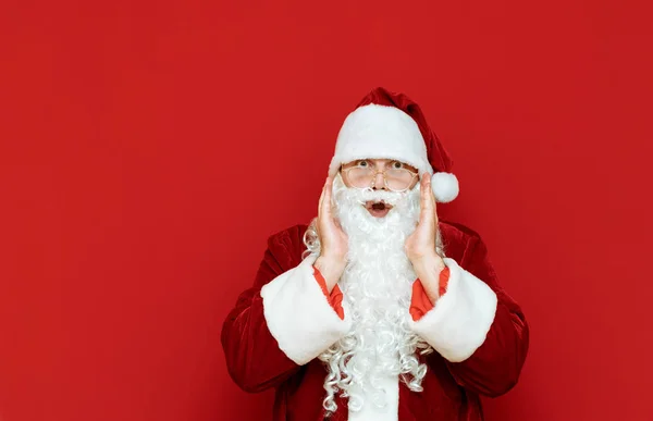 Asombroso Santa Claus se levanta contra una pared roja y mira a la cámara. Retrato del hombre sorprendido en traje de santa aislado. Santa se sorprendió de los descuentos para Navidad. Espacio de copia — Foto de Stock