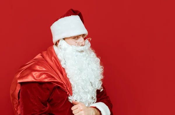 Arrodillarse Santa Claus con bolsa de regalos se encuentra en el fondo rojo y mira a la cámara, retrato de primer plano. Aislado. Un Santa astuto para Navidad. Concepto de X mas. Espacio de copia — Foto de Stock