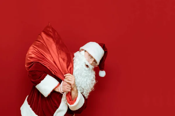 Funny Santa Claus lleva en su espalda una bolsa con regalos aislados sobre fondo rojo. Retrato de Santa llevando regalos a los niños. Santa y regalos para el concepto de Navidad. Año Nuevo. X mas — Foto de Stock