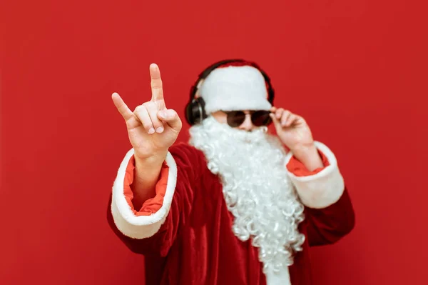 Santa Claus escucha música en auriculares sobre fondo rojo y muestra gesto Heavy Metal, aislado. El enfoque en la mano muestra el gesto de Rock. Santa Claus escucha música rock. Concepto X-mas . — Foto de Stock