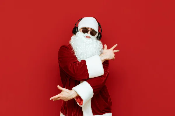 Retrato de estudio de Papá Noel alegre en gafas de sol escuchando música en auriculares y divirtiéndose sobre fondo rojo. Santa Claus fiestero con auriculares aislados sobre fondo rojo. X-mas — Foto de Stock