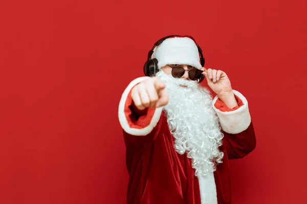 Papai Noel em óculos de sol e fones de ouvido escuta música, olha para a câmera e aponta o dedo para você. Conceito de Ano Novo e Natal . — Fotografia de Stock