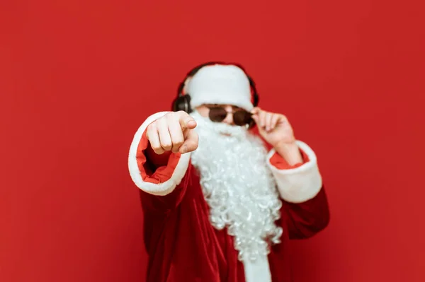 Santa Claus en gafas de sol y auriculares escucha música y señala con el dedo a la cámara sobre un fondo rojo. Concéntrate en el dedo índice que apunta hacia ti. Año Nuevo y concepto de Navidad . —  Fotos de Stock