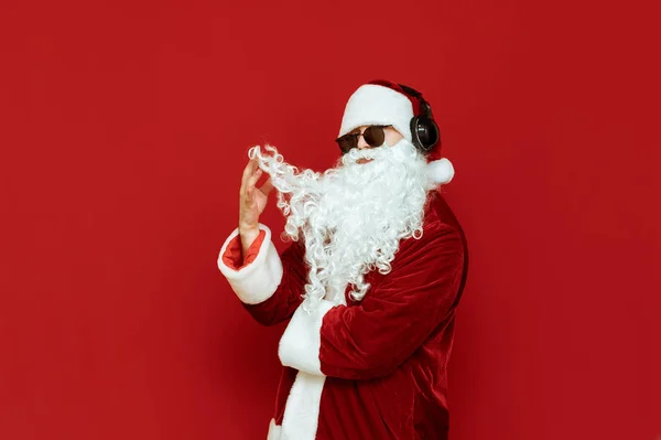 Studio zdjęcie modnego Świętego Mikołaja w okularach przeciwsłonecznych i słuchawkach stoi na czerwonym tle i bawi się brodą z poważną twarzą. Odizolowany portret pięknego Mikołaja. Koncepcja świąteczna. — Zdjęcie stockowe