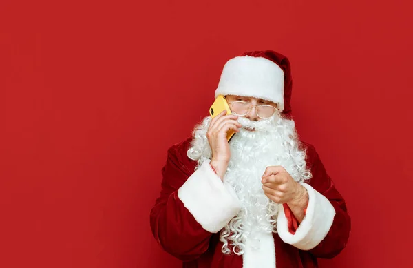 Retrato de un divertido Santa Claus hablando por teléfono sobre un fondo rojo y mirando hacia un lugar en blanco con una cara sospechosa.Saludo de Navidad en el teléfono desde Santa. Concepto de Año Nuevo — Foto de Stock