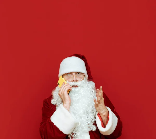 Impresionado Santa Claus hablando por teléfono sobre un fondo rojo y con una cara sorprendida mira a la cámara sobre un fondo rojo. Santa con smartphone y espacio en blanco para Copyspace — Foto de Stock