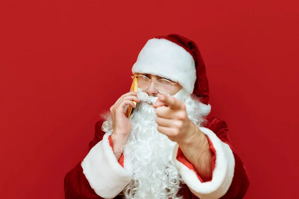 A close portrait of Santa Claus is ringing on the phone and pointing his index finger at the camera on a red background. Santa is calling you! Isolated. Christmas and New Year concept. Copyspace — Stock Photo, Image
