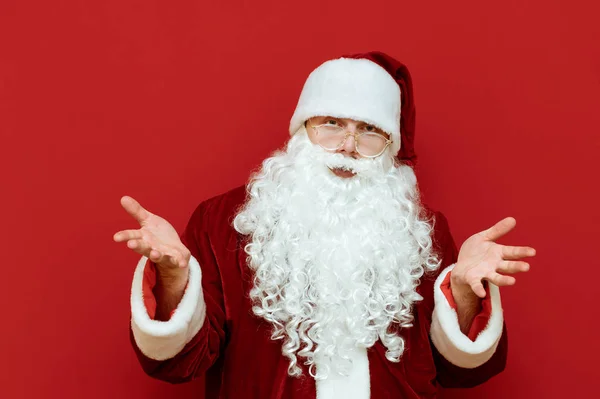 Das Porträt des verdutzten Weihnachtsmannes steht auf rotem Grund und breitet seine Hände seitlich aus. Lustiges Weihnachtsmann-Porträt mit traurigem Gesicht, isoliert. Neujahrskonzept. — Stockfoto