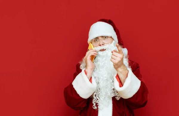 Portret Świętego Mikołaja rozmawiającego przez telefon i aktywnie gestykulującego na czerwonym tle, spoglądającego w kamerę z poważną twarzą. Mikołaj wita ludzi przez telefon i wskazuje kamerą — Zdjęcie stockowe