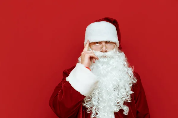 Das Porträt des Weihnachtsmannes zeigt Gesten des Nachdenkens, hält sich den Finger an den Kopf und blickt mit ernstem Gesicht in die Kamera. nachdenklicher Weihnachtsmann isoliert auf rotem Hintergrund. Weihnachtskonzept. Weihnachten — Stockfoto