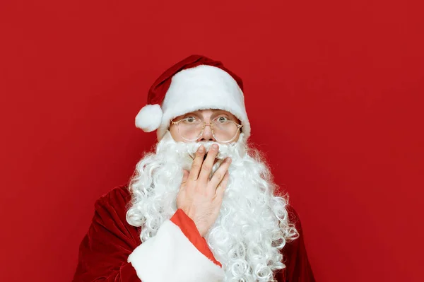 Primer plano retrato de estudio de Santa Claus conmocionado sobre fondo rojo, se cubrió la boca con asombro, mira a la cámara con la cara asombrada. Emocional Santa aislado sobre fondo rojo.Concierto de Navidad — Foto de Stock