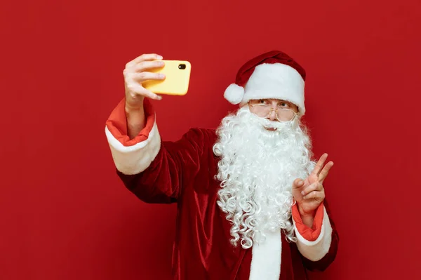 Studiofoto von Weihnachtsmann, der auf Smartphone-Kamera posiert, Selfies macht und vor rotem Hintergrund Friedensgeste an -geste zeigt. Selfies. Weihnachtsmann und Technologie. Frohe Weihnachten. — Stockfoto