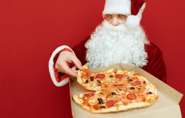 Elinde bir kutu pizza olan Noel Baba kırmızı arka planda duruyor, lezzetli bir dilim sıcak pizza alıyor ve kameraya bakıyor, eline bir parça pizza ile odaklanıyor. Noel Baba pizza yiyor, izole edilmiş.