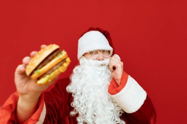 Pozitif Noel Baba elinde hamburgerle kırmızı arka planda duruyor, kameraya bakıyor ve gülümsüyor. Noel Baba fast food sever. İzole edilmiş. Noel konsepti.