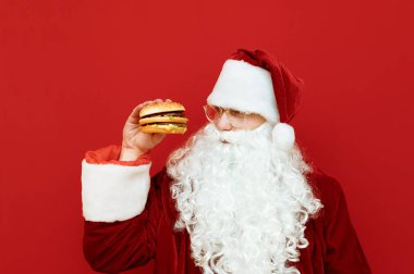 Elinde hamburgerle kırmızı arka planda duran, aç ve fast food arayan Noel Baba kostümlü bir adam portresi. Noel Baba fast food yiyecek. Kırmızı arka planda izole edilmiş. Boşluğu kopyala