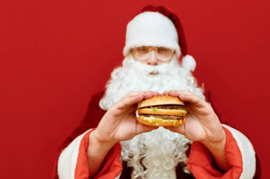 Noel Baba 'nın elinde kırmızı arka planda izole edilmiş lezzetli bir burger var. Noel Baba' nın elinde sakallı ve gözlüklü burger kırmızı arka planda duruyor ve fast food arıyor. Noel konseptinde fast food.