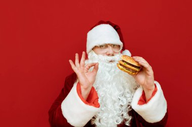 Neşeli Noel Baba elinde hamburgerle kırmızı arka planda duruyor, tamam işaretini gösteriyor, kameraya bakıyor ve gülümsüyor. Noel Baba kılığındaki adam fast food seviyor. Zararlı yiyecekler. Noel