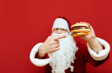 Noel Baba elinde bir hamburger tutuyor ve parmağını fast food 'a doğrultuyor. Bir adamın elindeki hamburgere odaklanıyor. Noel Baba fast food yemeyi sever. Yeni yıl ve Noel. Geçmiş. Kopyalama alanı