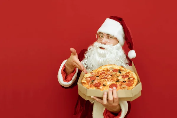 Yakışıklı Noel Baba pizza dağıtıcısı elinde pizza kutusuyla kırmızı arka planda izole edilmiş elini lezzetli parçalar halinde gösteriyor ve kameraya bakıyor. Noel Baba Noel için pizza getiriyor..