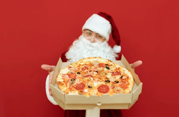 Santa Claus hält eine Schachtel mit heißer leckerer Pizza in der Hand und zeigt sie der Kamera. Box mit frischer Pizzalieferung in der Hand in Weihnachtsmann. Pizza zu Weihnachten. Neues Jahr mit Pizza. — Stockfoto