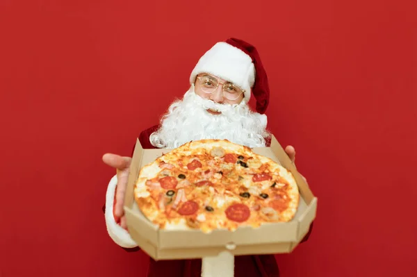 Lächelnd steht Weihnachtsmann Claus vor rotem Hintergrund, in ihren Händen hält sie eine Schachtel leckere heiße Pizza und richtet sie in die Kamera. Weihnachtsmann liefert Pizza zu Weihnachten, blickt lächelnd in die Kamera — Stockfoto