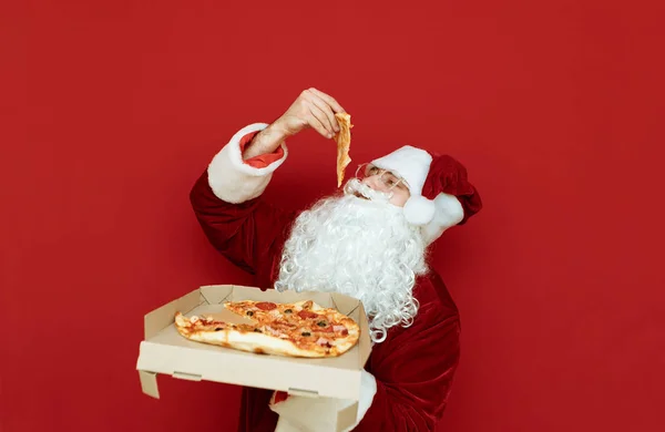 Noel Baba kostümü giymiş aç bir adam elinde pizza kutusuyla kırmızı arka planda duruyor ve bir parça yiyor. Noel Baba pizza yemeyi sever. Noel konsepti. Boşluğu kopyala