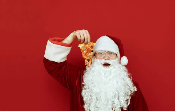 Portrait of shocked Santa Claus standing on red background with a slice of delicious pizza in hand with shocked face looking in camera. Empty space for text on the left.Discount pizza for Christmas