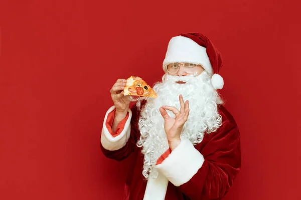 Facet w kostiumie Mikołaja je pizzę. Pizza na Święta. Przystojny Mikołaj z kawałkiem pizzy w ręku stoi na czerwonym tle, patrzy w kamerę i pokazuje znak kciuka Ok. Copyspace — Zdjęcie stockowe