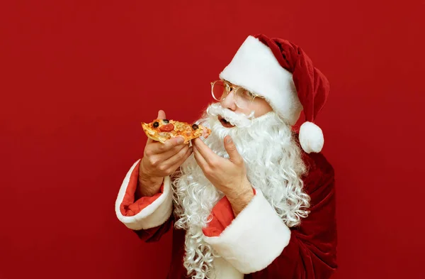 Zabawny Mikołaj jedząc pyszną pizzę na czerwonym tle i patrząc w aparat. Mikołaj uwielbia jeść pizzę na święta, trzymając kawałek, odizolowany. Santa Claus je Fast Food. — Zdjęcie stockowe