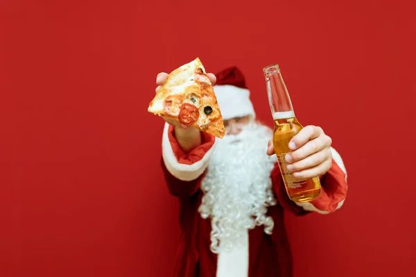 Święty Mikołaj pokazuje w aparacie butelkę piwa i kawałek pizzy na czerwonym tle. Pizza i butelka w rękach Mikołaja odizolowane na czerwonym tle. Koncepcja świąteczna. — Zdjęcie stockowe