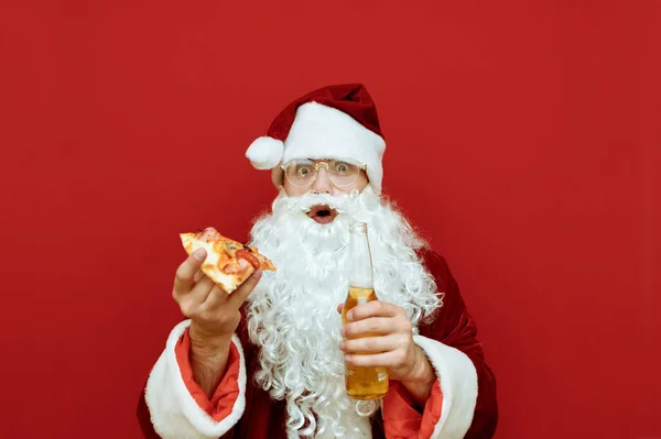 Zaskoczony Święty Mikołaj z kawałkiem pizzy i butelką piwa w ręku stoi na czerwonym tle, patrzy w kamerę z zaszokowaną twarzą, odizolowany. Śmieszny Mikołaj je śmieciowe jedzenie i pije alkohol. — Zdjęcie stockowe