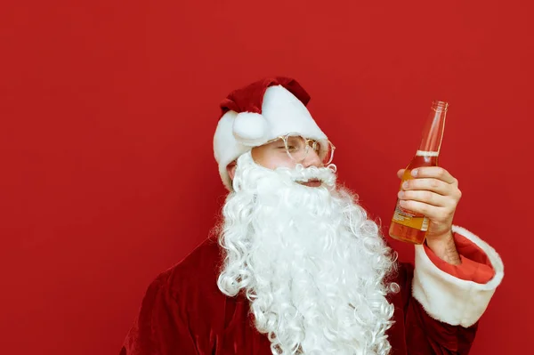 A funny Santa Claus portrait wearing glasses and a beard stands on a red background with a beer bottle in his hand and looks away at alcohol. Bad Santa drinks beer. Copy space — Stock Photo, Image