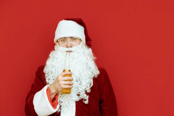 Portrait of drunk Santa Claus with bottle of beer in hands on red background, looks into camera. Santa in alcoholic span isolated on red background. Drunk man santa for new year. — Stock Photo, Image