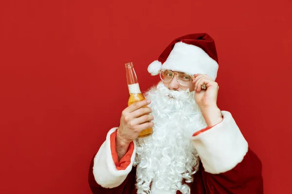 Zbliżenie portret wesołego Mikołaja z butelką piwa w ręku, dopasowuje okulary, patrzy w kamerę i uśmiecha się. Śmieszny Mikołaj pije alkohol i pozuje przed kamerą. — Zdjęcie stockowe