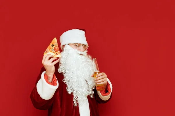 Szczęśliwy Mikołaj z butelką piwa i kawałkiem pizzy w rękach stoi na czerwonym tle, patrzy w kamerę i uśmiecha się. Mikołaj pije alkohol i je fast food. Boże Narodzenie. — Zdjęcie stockowe