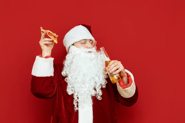 Radosny Święty Mikołaj z kawałkiem pizzy i butelką piwa w ręku stoi na czerwonym tle z zamkniętymi oczami, uśmiechnięty i dobrze się bawiący. Zabawny człowiek w stroju Mikołaja bawiący się, odizolowany. — Zdjęcie stockowe
