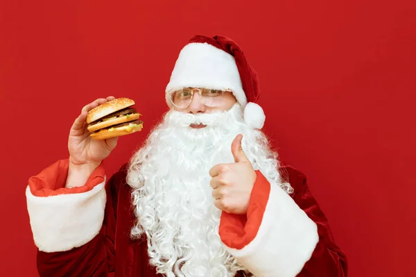 Portret szczęśliwego Mikołaja na czerwonym tle z hamburgerem w ręku, spogląda w kamerę, pokazuje kciuk w górę i uśmiecha się. Mikołaj uwielbia jeść fast food, trzyma kanapkę i pozuje do kamery. Boże Narodzenie — Zdjęcie stockowe