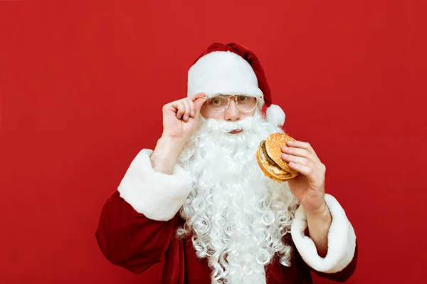 하얀 수염 과 안경을 쓰고 빨간 배경에 서 있는 산타 클로 오스 는 카메라 와 미소를 지으면서 버거 를 손에 들고 있다. 손에 햄버거를 들고 빨간 배경으로 격리 된 산타클로스 의사 진 — 스톡 사진