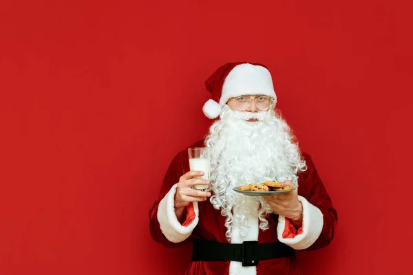 귀여운 산타클로스가 우유 한 잔 과 초콜릿 쿠키 한 접시를 들고 빨간 배경에 서서 카메라를 들여다보며 미소를 짓습니다. 크리스마스와 새해 컨셉이야. 고립되어 있습니다. Copyspace — 스톡 사진