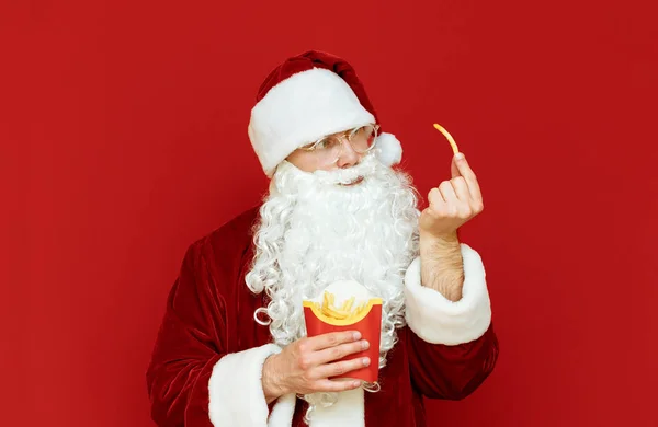 Portret zabawnego Mikołaja z frytkami w dłoniach na czerwonym tle. Mikołaj jedzący fast food na czerwonym tle. Przestrzeń kopiowania — Zdjęcie stockowe