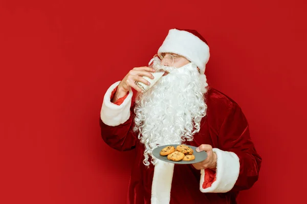 Święty Mikołaj pije mleko ze szklanki i trzyma w ręku talerz czekoladowych ciasteczek, odwracając wzrok od pustej przestrzeni. Mężczyzna w stroju Mikołaja pije mleko i je ciasteczka. Koncepcja nowego roku. — Zdjęcie stockowe