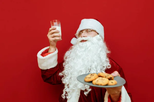 Portret szczęśliwego Mikołaja na czerwonym tle, trzymającego szklankę mleka i talerza z ciasteczkami, uśmiechającego się i patrzącego w kamerę. Mikołaj podniósł szklankę mleka i raduje się. Boże Narodzenie i Nowy Rok. — Zdjęcie stockowe