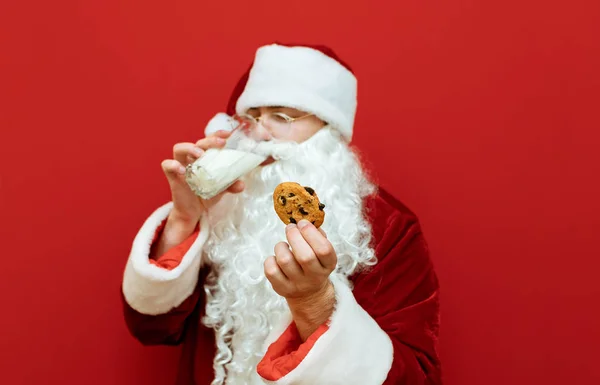 Le Père Noël boit du lait et tient des biscuits de Noël dans sa main. Concentrez-vous sur les biscuits de Noël entre les mains du Père Noël. Contexte. Isolé sur fond rouge. Concept Nouvel An . — Photo