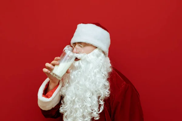 Portret z bliska Świętego Mikołaja pijącego mleko ze szklanki na czerwonym tle i odwrócił się.Mężczyzna w stroju Mikołaja pijącego mleko, odizolowany. Koncepcja świąteczna. — Zdjęcie stockowe