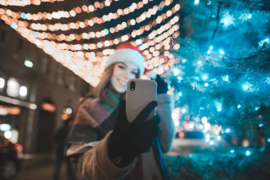 Akıllı telefona odaklan. Noel Baba şapkalı mutlu kızın portresi Noel ışıklarıyla süslenmiş sokak arkaplanındaki Noel ağacının yakınındaki akıllı telefondan selfie çekiyor..