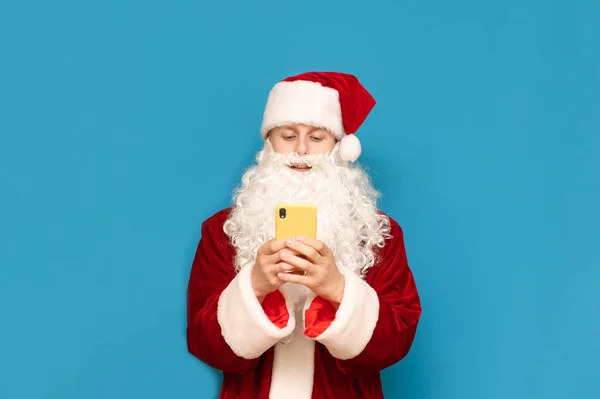Leende ung man i tomte kostym stående på blå bakgrund och med hjälp av smartphone, isolerade. Santa tonåring med smartphone isolerad på blå bakgrund. Julkoncept. — Stockfoto