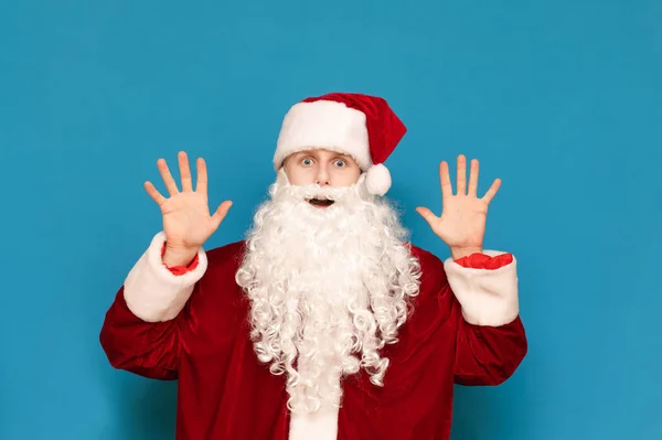 Retrato de un joven Santa asustado sobre un fondo azul, levantado de las manos conmocionadas y mirando a la cámara con una cara emocional. Chico sorprendido con disfraz de santa aislado. Concepto de Navidad . — Foto de Stock