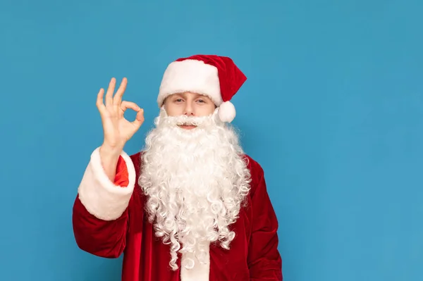 Der fröhliche junge Weihnachtsmann mit Bart und rotem Hut steht auf blauem Grund, zeigt seine Fingergeste und blickt in die Kamera. Kerl im Weihnachtsmann-Anzug zeigt das Zeichen ok und lächelt. — Stockfoto