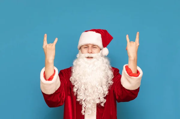 Młody Santa Claus rocker stoi na niebieskim tle i pokazuje rękami znak Heavy Metal patrząc w kamerę.Santa słucha muzyki rockowej, pokazuje znak rock.Portret rock and rolla Santa — Zdjęcie stockowe