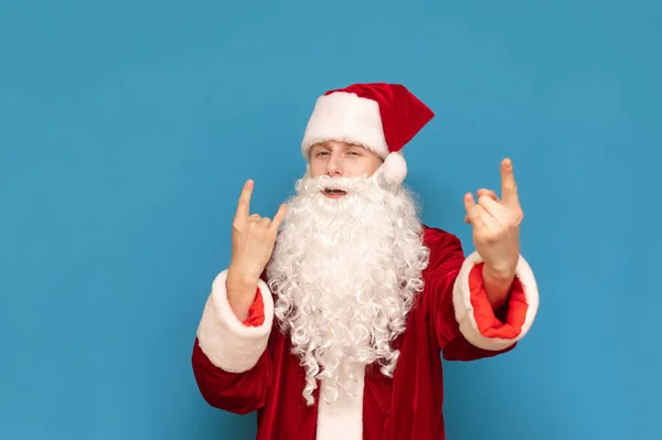 Веселый юный Санта Клаус показывает металлический знак и смотрит в камеру, парень в костюме Санты любит слушать рок-музыку. Playful Santa показывает рок-жест и позирует на камеру . — стоковое фото
